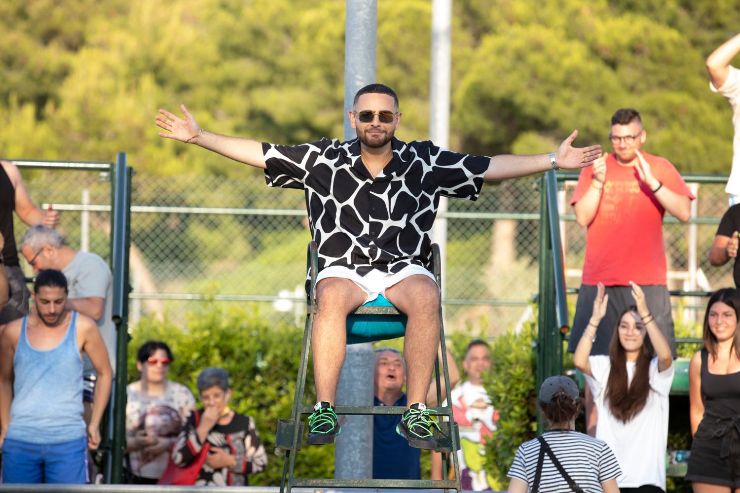 Un uomo con camicia a macchie bianche e nere siede sul seggiolone dell'arbitro a braccia aperte, con il pubblico dietro