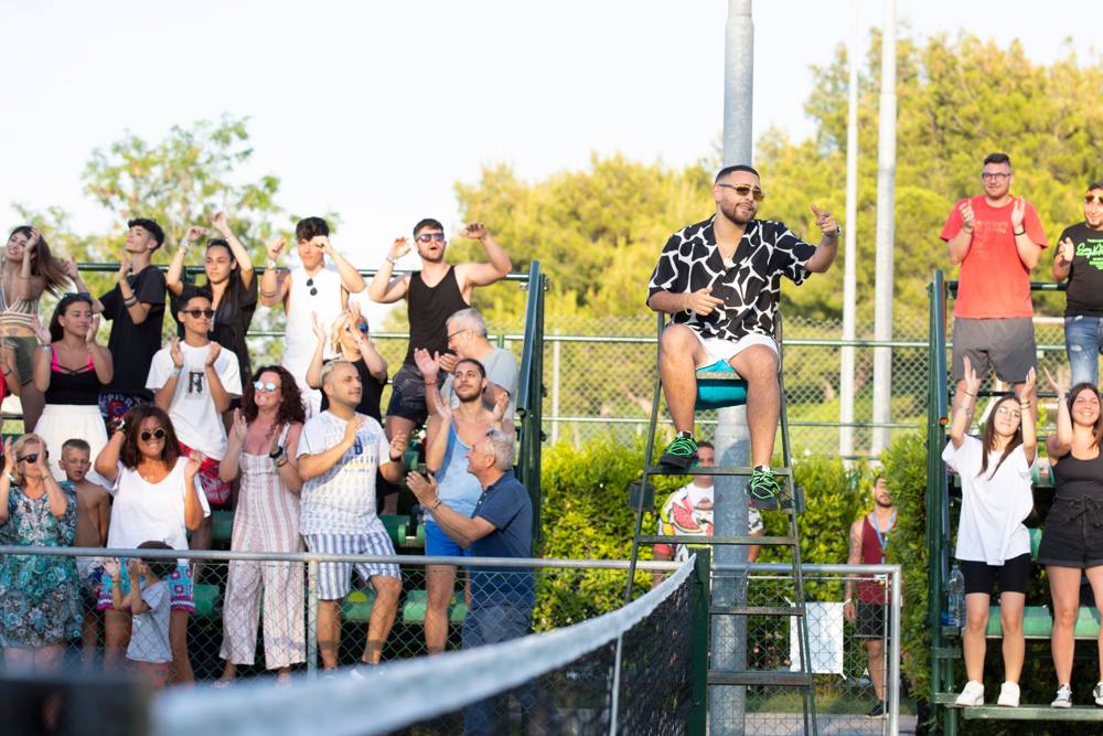 L'uomo in camicia maculata sul seggiolone indica verso l'obiettivo mentre il pubblico in tribuna applaude e balla