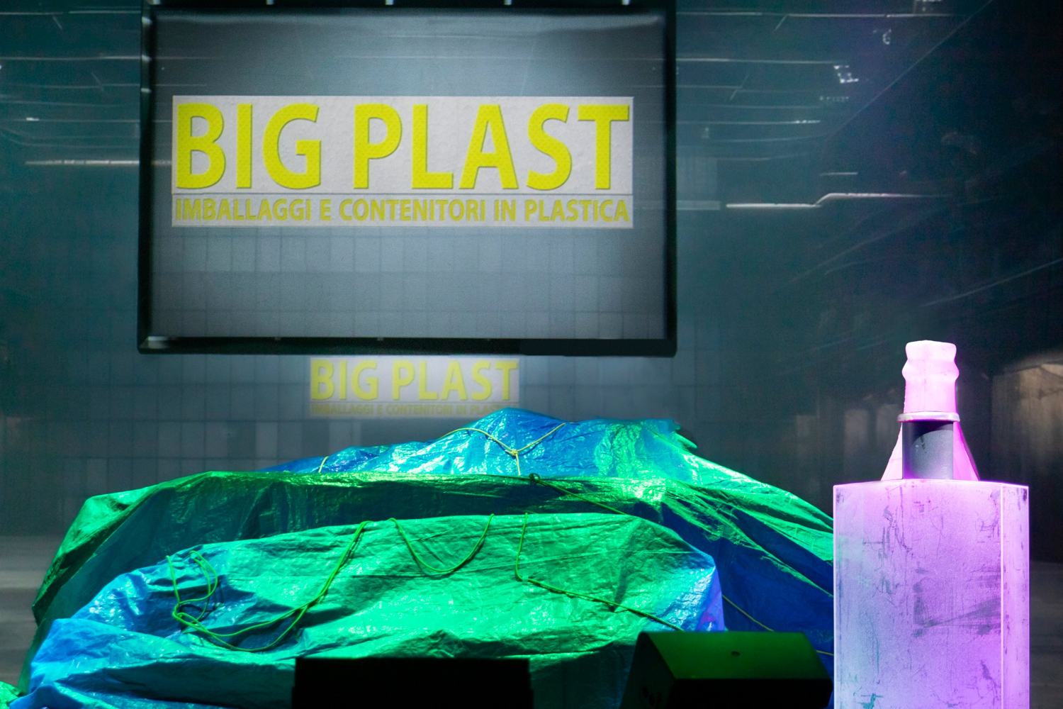 Uno schermo con la scritta Big Plast, imballaggi e contenitori in plastica, sopra un telo verde e blu legato con corde; a destra una bottiglia rosa su un piedistallo