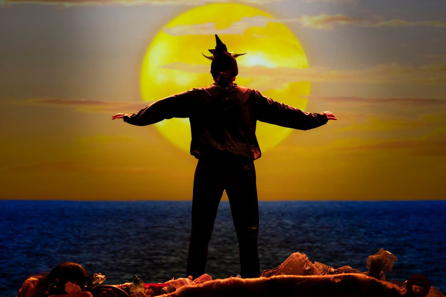Una figura in silhouette a braccia aperte davanti a un enorme sole giallo che tramonta sul mare
