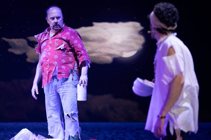Un uomo in camicia rosa a fiori strappata guarda un altro uomo di spalle, vestito di bianco, davanti a una nuvola illuminata dalla luna
