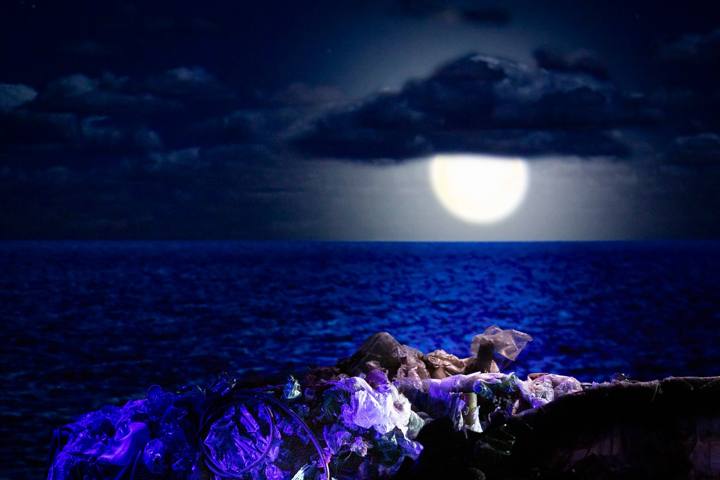 Luna piena su un mare blu notte, in primo piano una montagna di rifiuti di plastica illuminata di viola