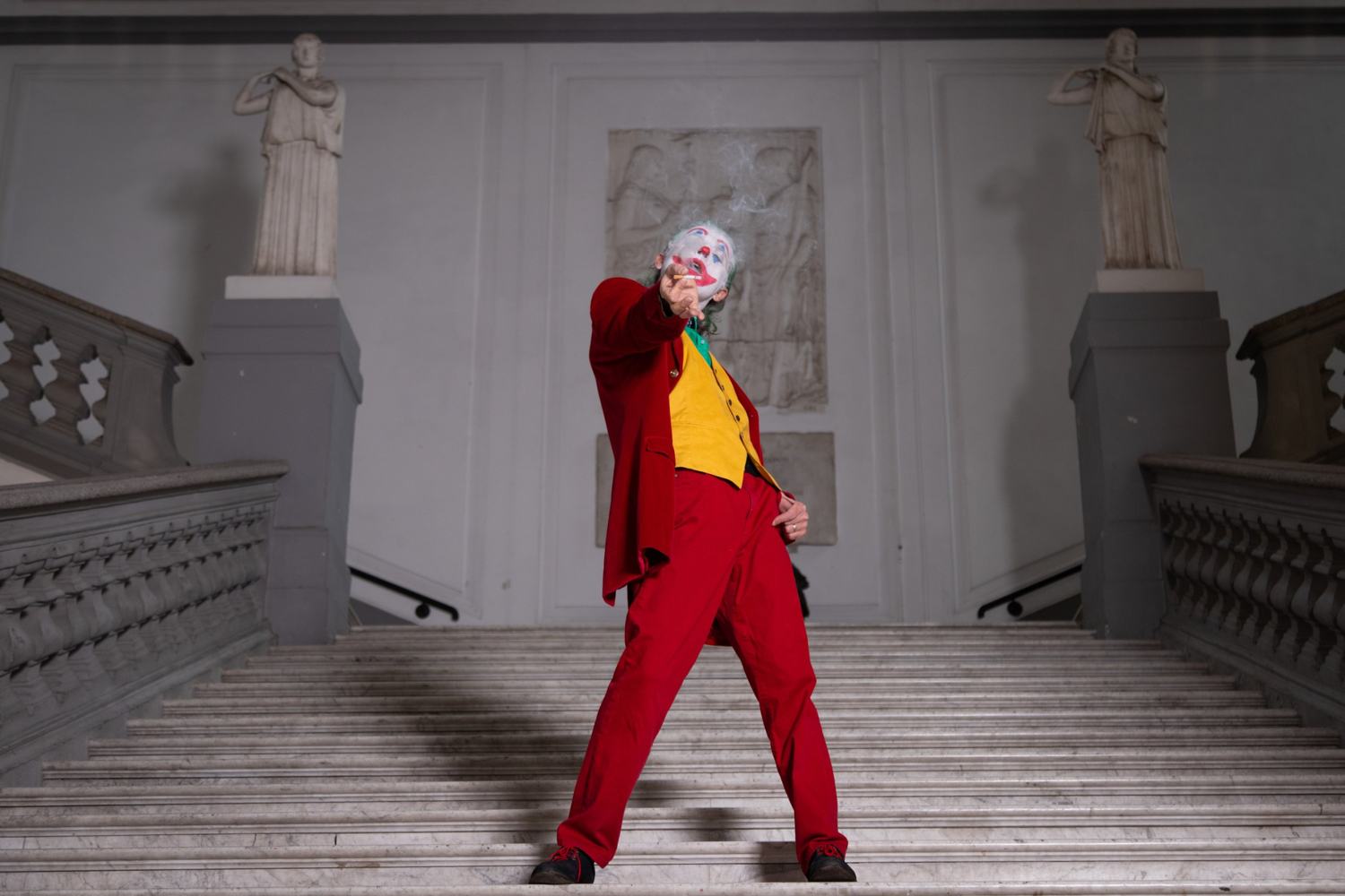 Joker in basso sulla scalinata di un palazzo, tra due statue, punta la sigaretta verso l'obiettivo