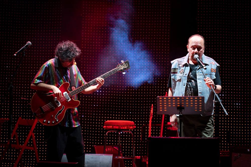 Un bassista con camicia a paillettes e un uomo con gilet di jeans che canta al microfono, davanti a una parete di led rossi