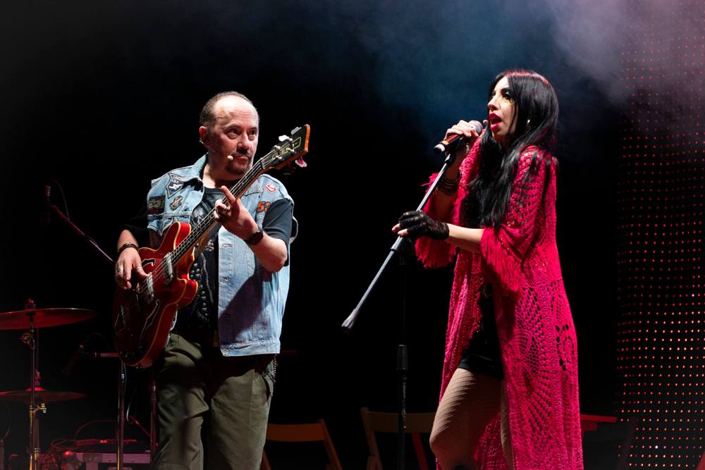 Un uomo con gilet di jeans suona il basso e guarda una cantante con lunga veste rosa che canta al microfono nel fumo