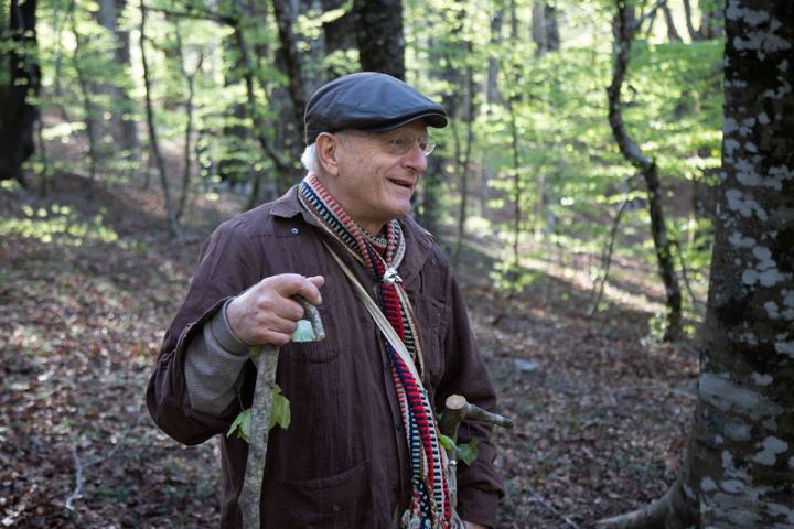 Ritratto di un uomo anziano con coppola, occhiali e sciarpa a righe, che regge un bastone di legno e guarda lontano nel bosco